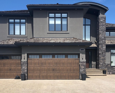 Modern two-story house in Edmonton with three wooden garage doors featuring decorative windows and stone accents around the entrance and garage area, showcasing quality garage door service.