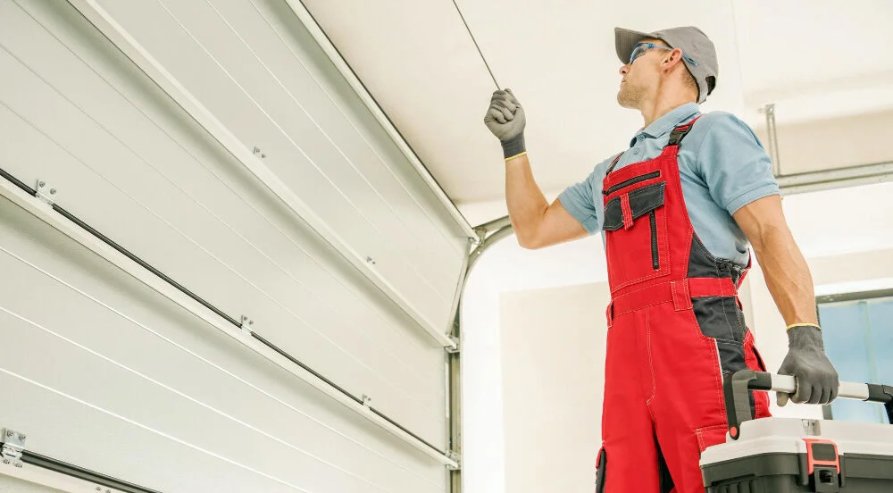 Technician In Overalls Inspecting A Garage Door Spring Inside A Residential Garage, Holding A Toolbox